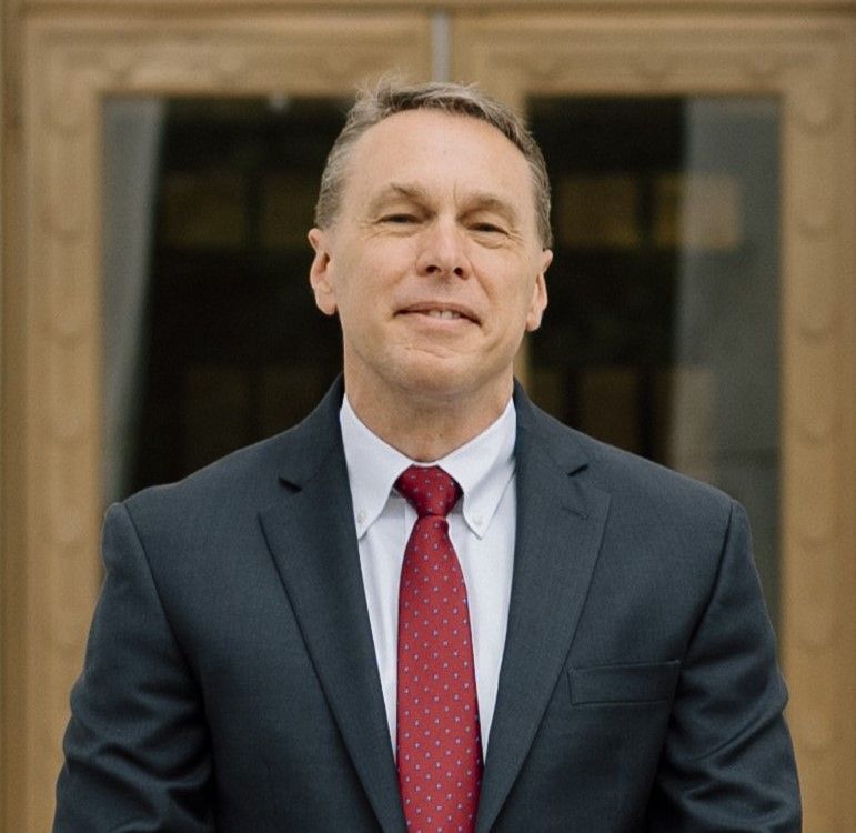 Man in suit and red tie stands outdoors, smiling slightly, in front of a building entrance.