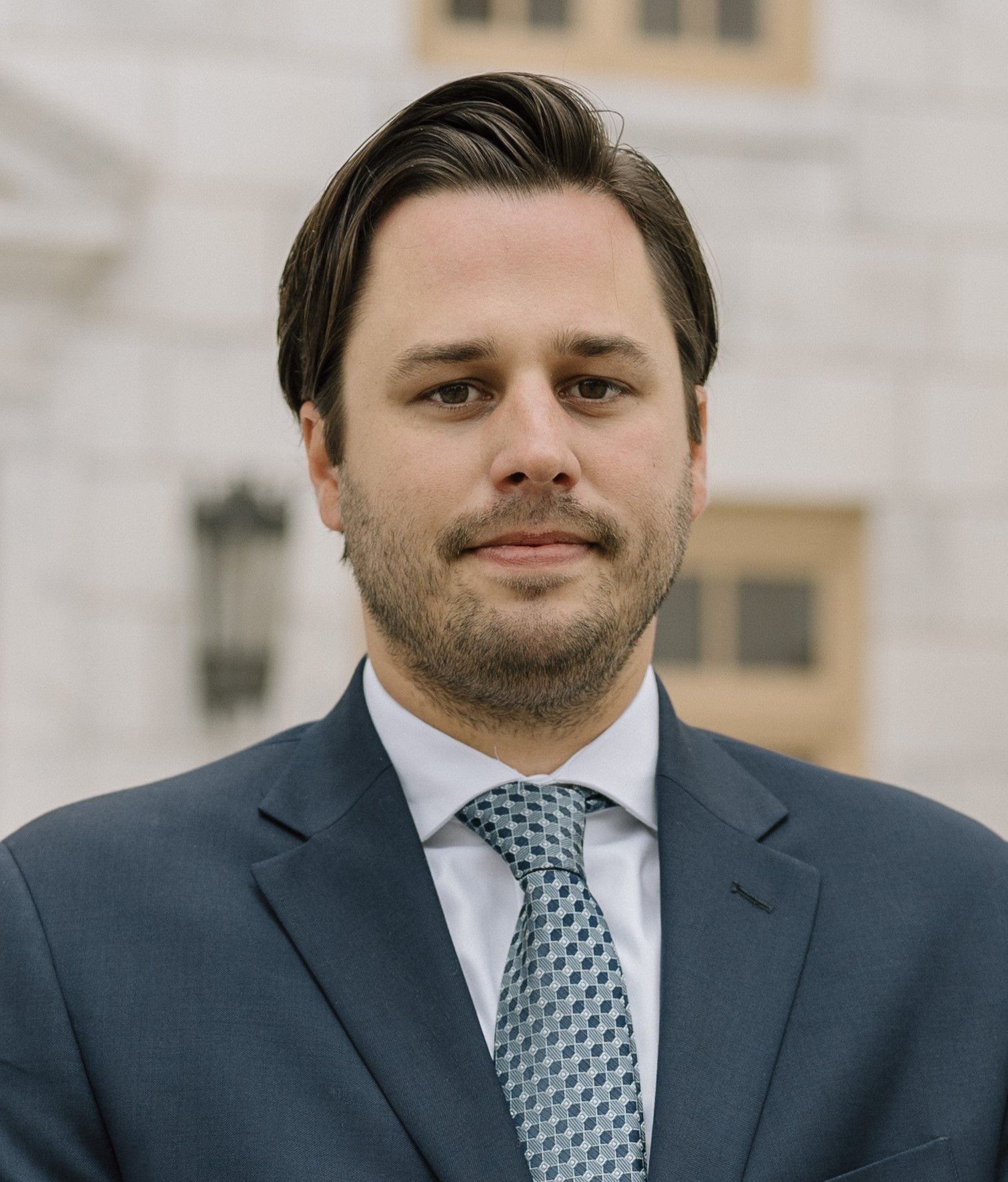 Man in a suit and tie, posing in front of a light-colored building.