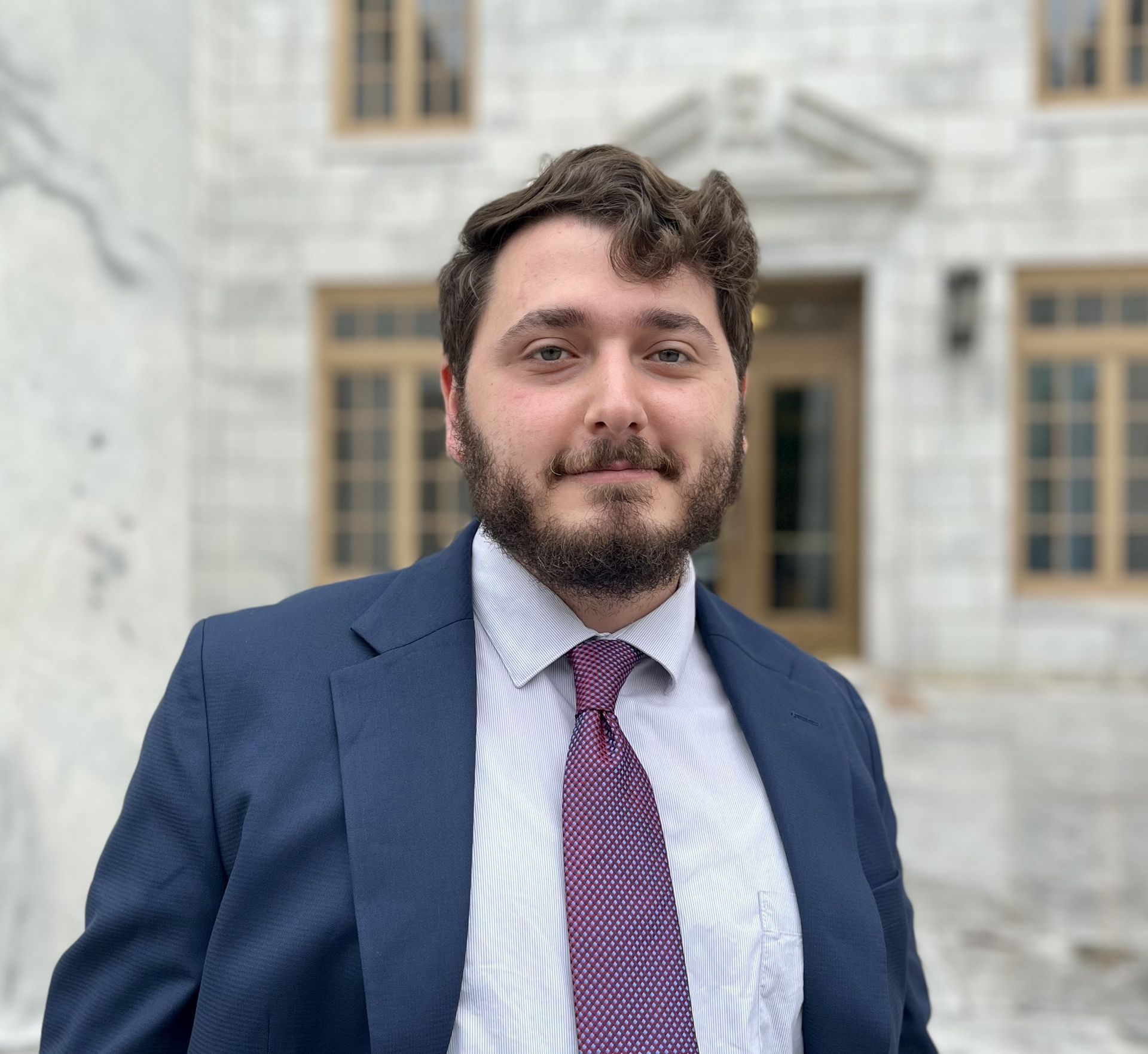 Man in a blue suit and purple tie stands in front of a light-colored building.