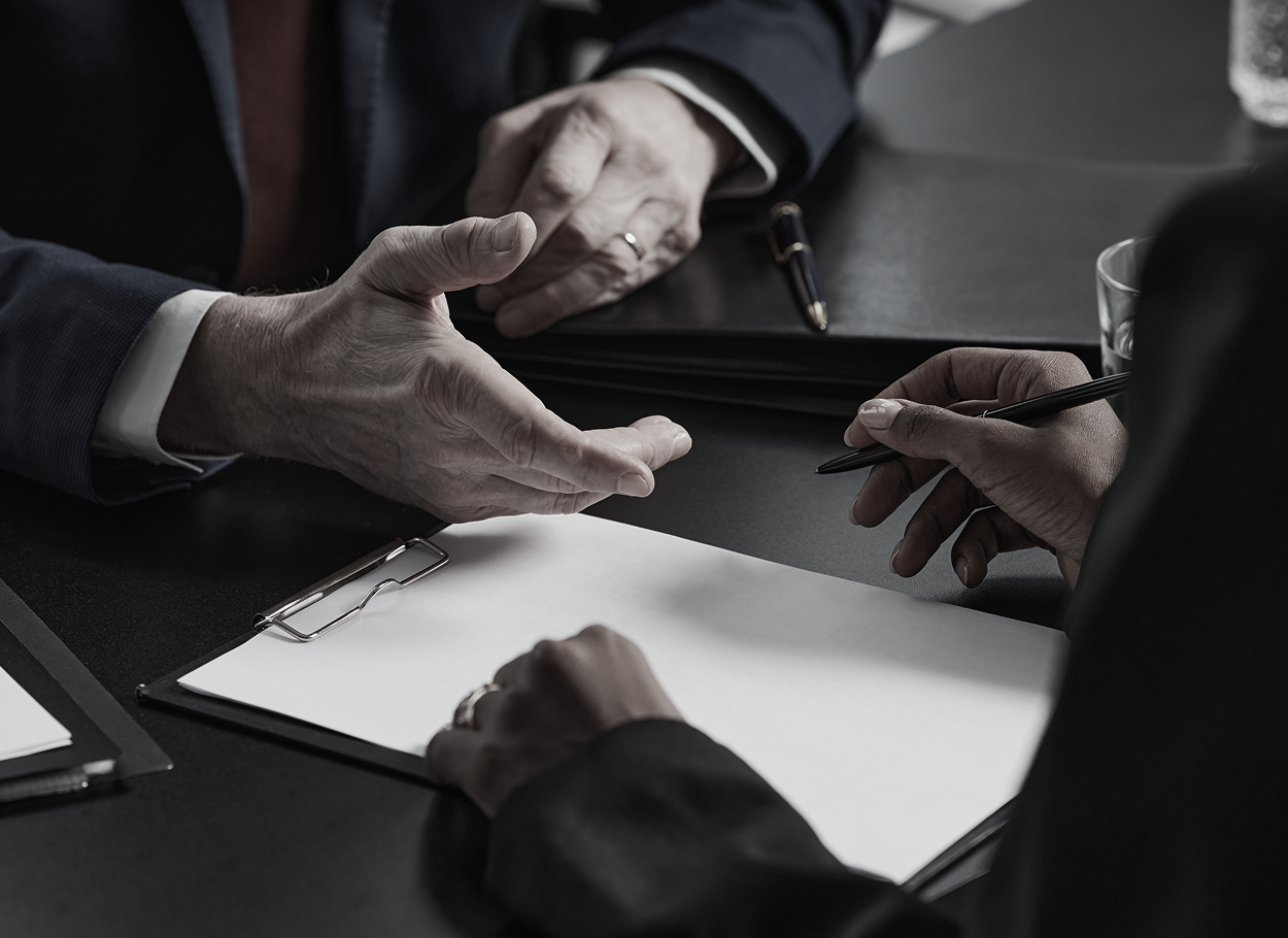 Two people at a table, one gesturing with hands, the other writing on a document.