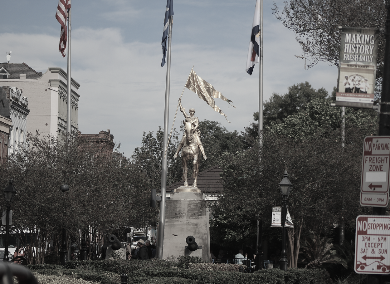 Statue of a man on horseback holding a banner in a city square, surrounded by flags and trees.