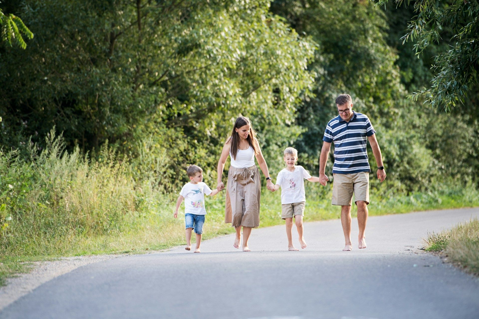 Family of four walking barefoot on a paved path in a park, holding hands.