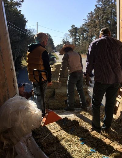 Three people stacking hay bales inside a barn with a road visible outside.