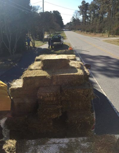 Hay bales stacked on a flatbed truck near a road, with workers in the background. Sunny day.