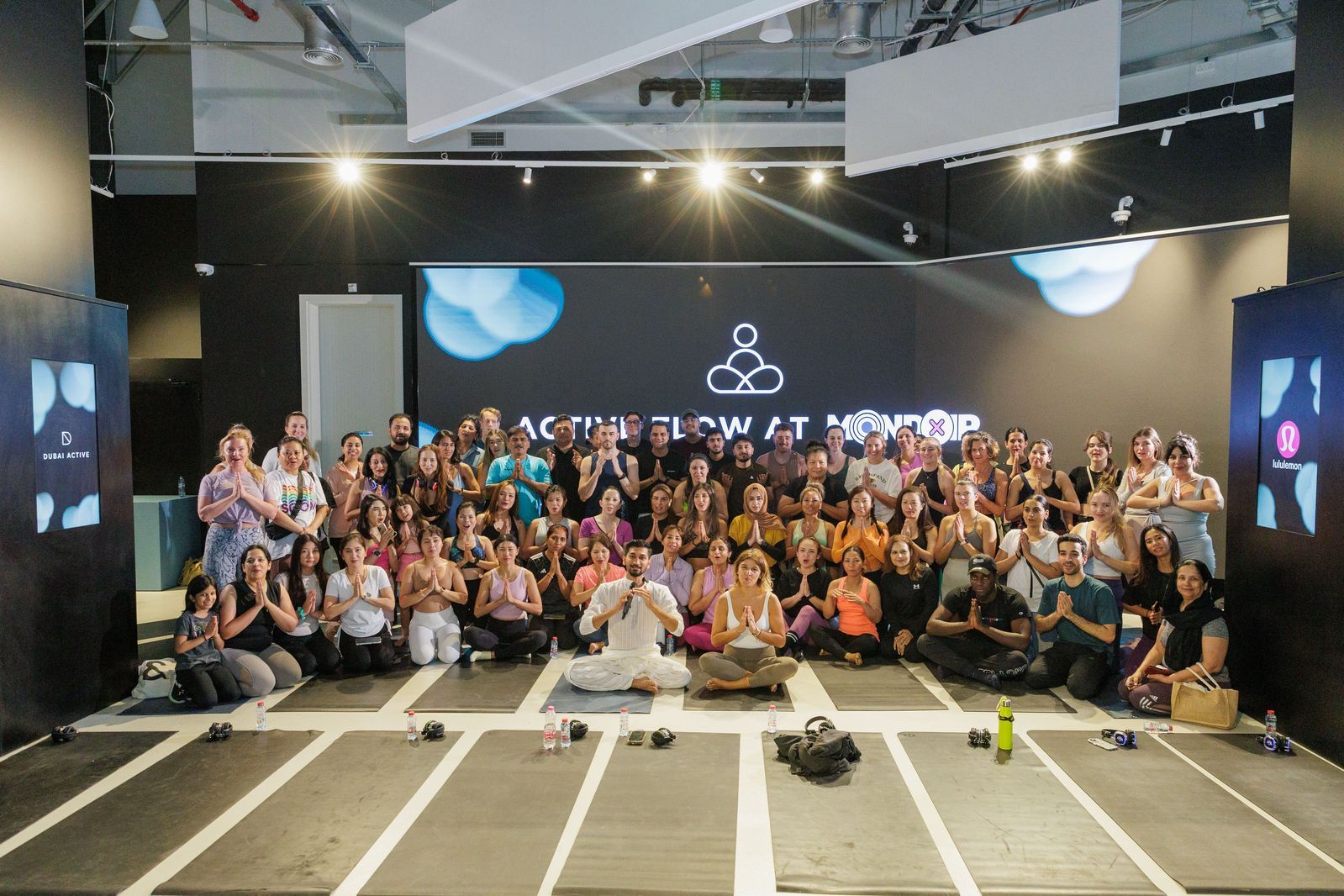 A large group of people are sitting on yoga mats in front of a large screen.