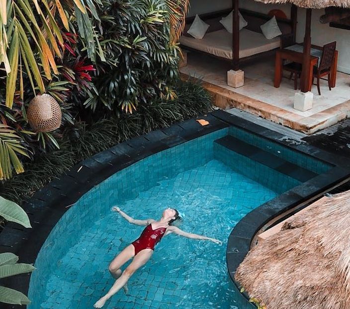 Woman in red swimsuit floats in a blue pool. Tropical setting with daybed and thatch roof.