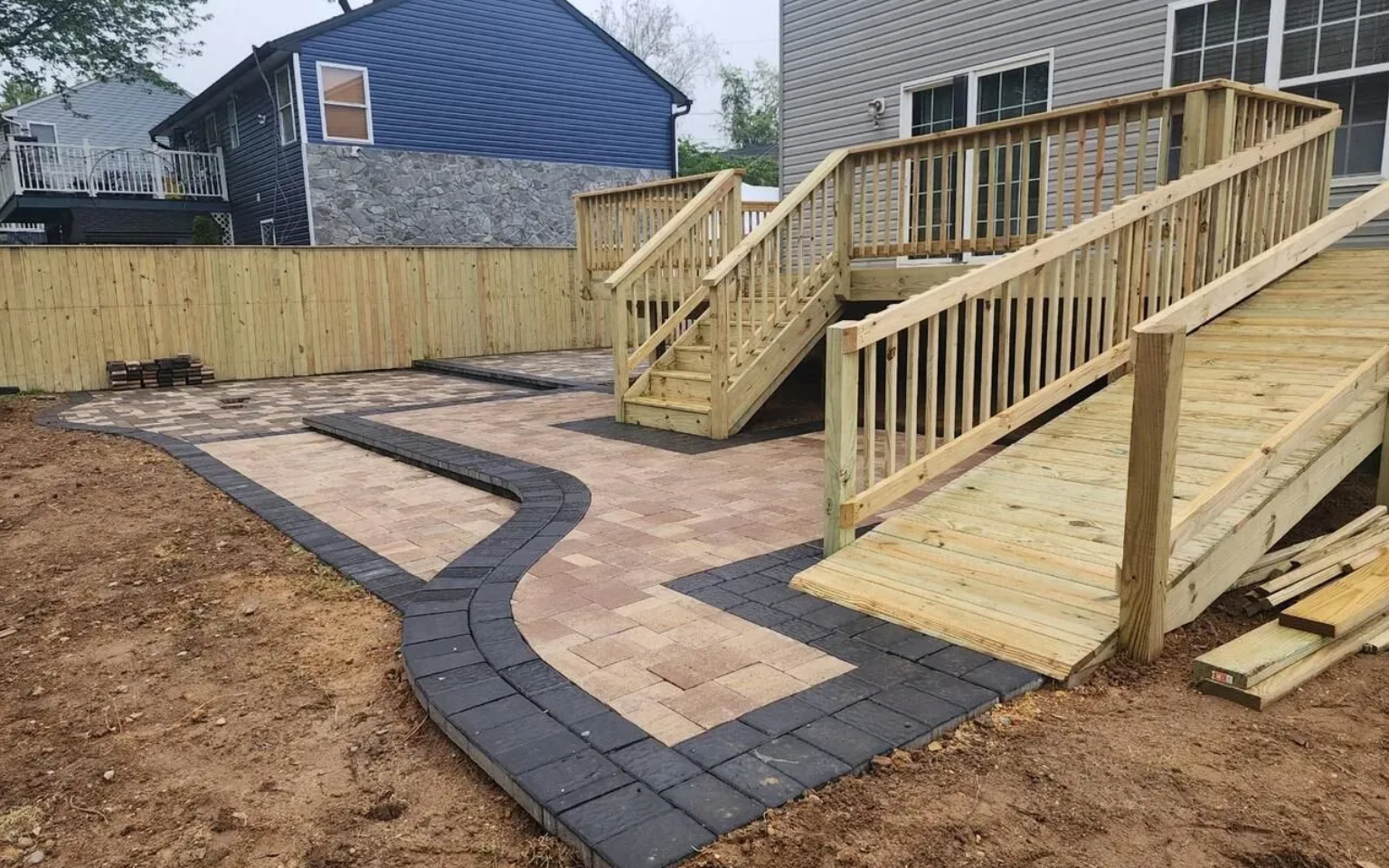A wooden deck with stairs and a ramp leading to a brick patio in a backyard setting.