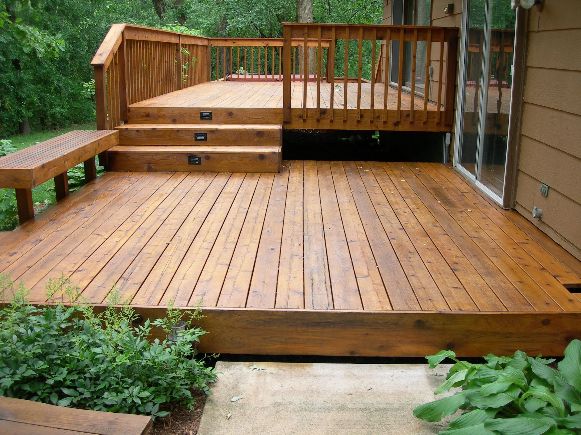 Wooden deck with steps, a bench, and a railing next to a house with sliding glass doors.