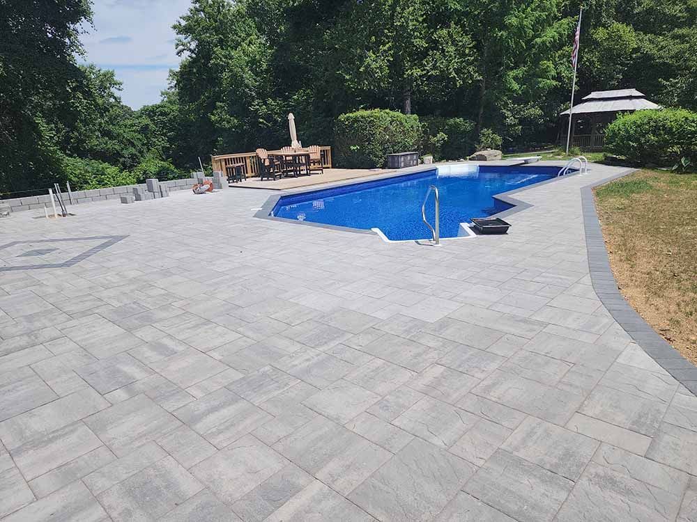A large swimming pool is surrounded by a brick patio and a white fence.