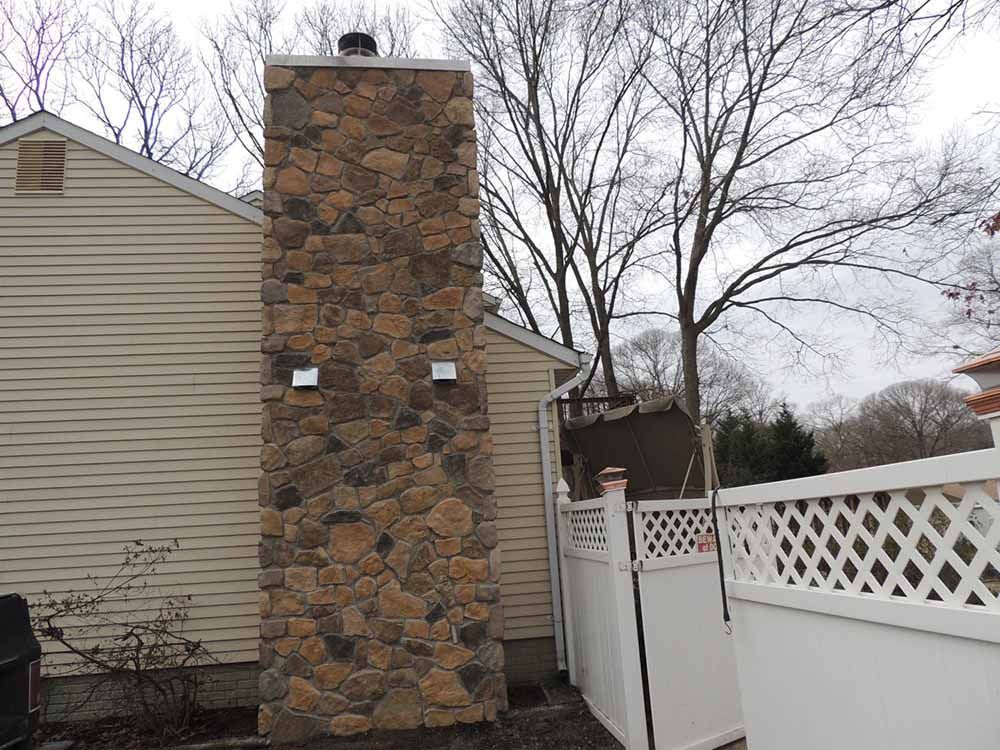 A large stone chimney on the side of a house next to a white fence.