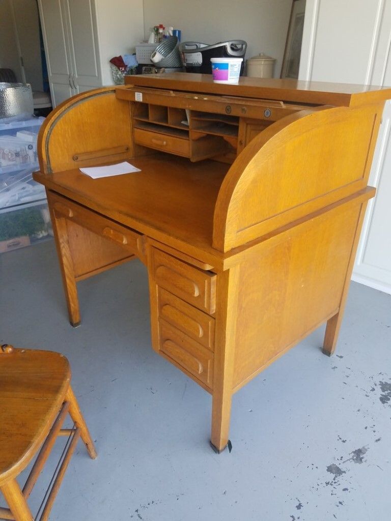 A wooden roll top desk is sitting in a garage next to a chair.