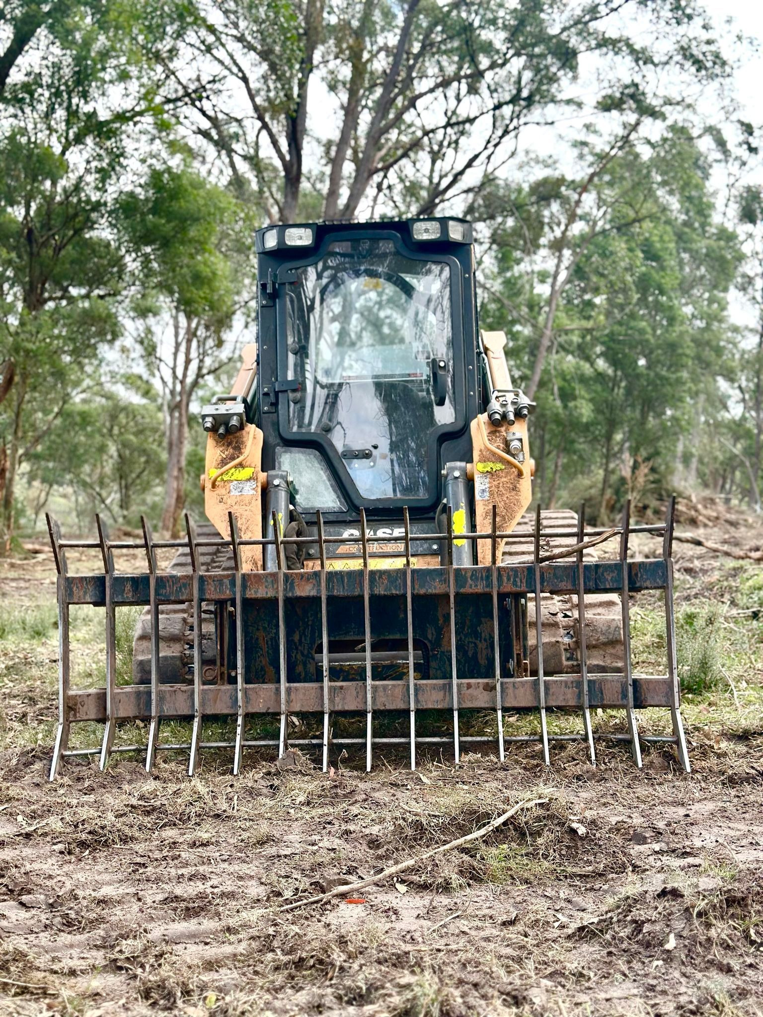 A yellow skid steer with a rake attachment clearing debris from a muddy field, with trees in the background.