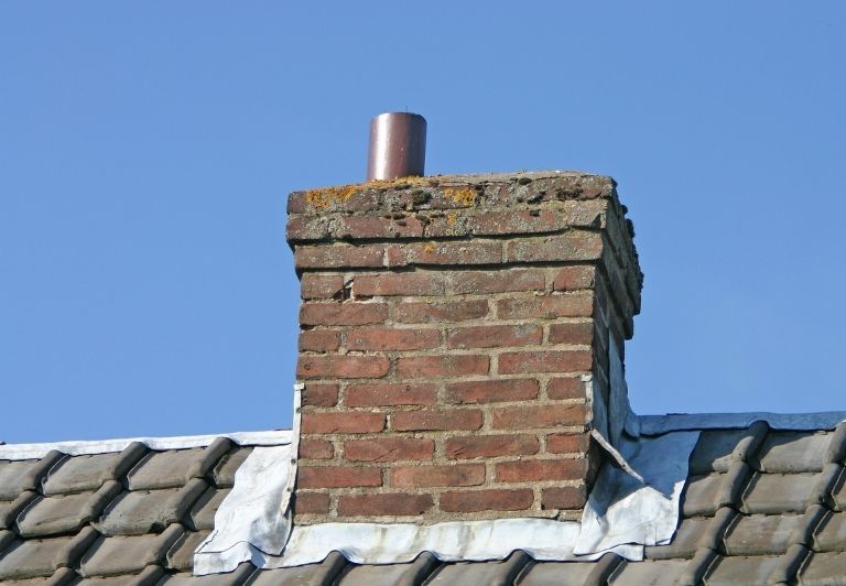 Chimney sweep on a roof, wearing safety harness, cleaning a brick chimney against a blue sky.