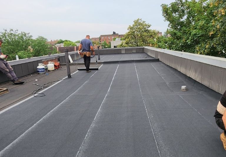 Person applying black sealant to a wood surface with a roller.  A paint tray filled with sealant is nearby.