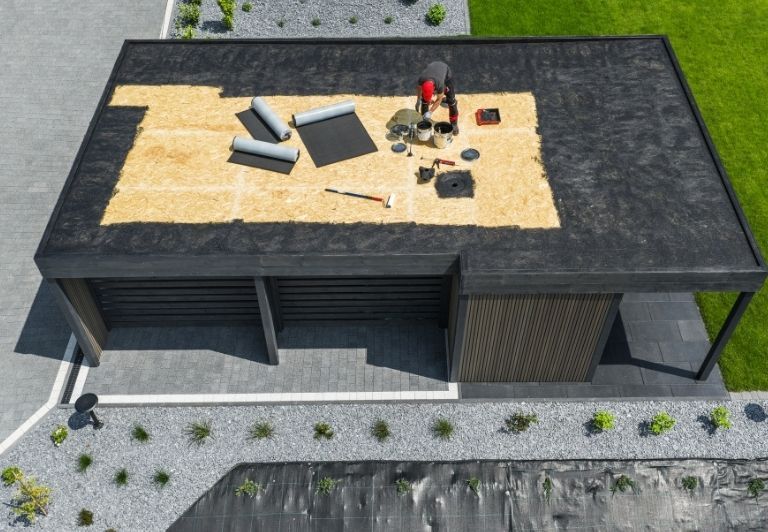 Roofers working on a flat roof, applying roofing material. Black roof, blue sky, trees in the background.