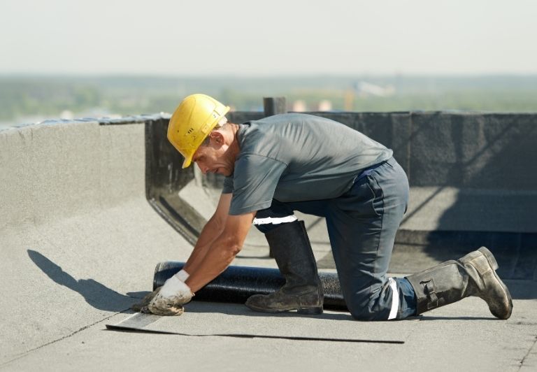 Workers installing a roofing membrane, using a torch. They wear blue work clothes, boots, and gloves, working on a flat roof.