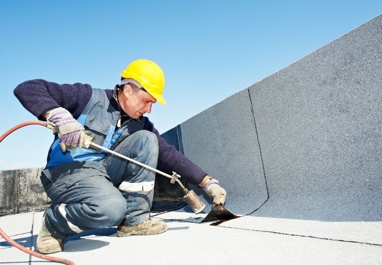 Flat, gray commercial rooftop with various pipes and equipment. Scaffolding visible on the side, trees in the background.