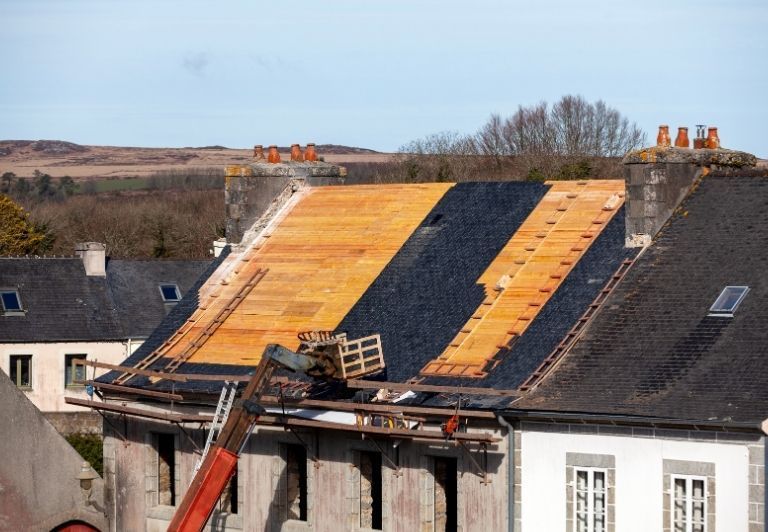 Brick building under construction, roof partially tiled with red and green tiles, scaffolding visible.
