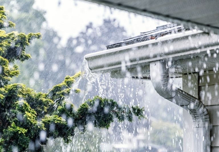 Person installing a black rain gutter on a roof, wooden scaffolding visible.