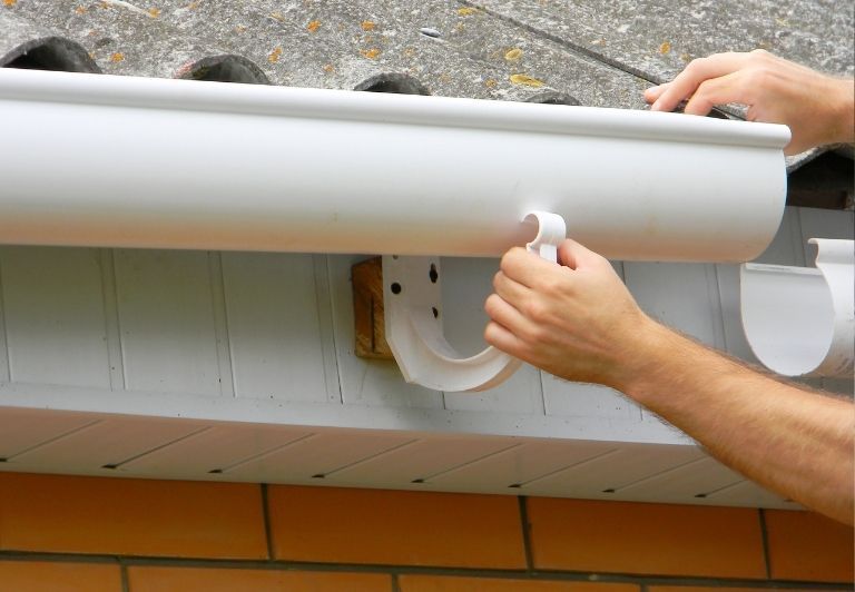 Person installing a white gutter on a roof, using a ladder for access.
