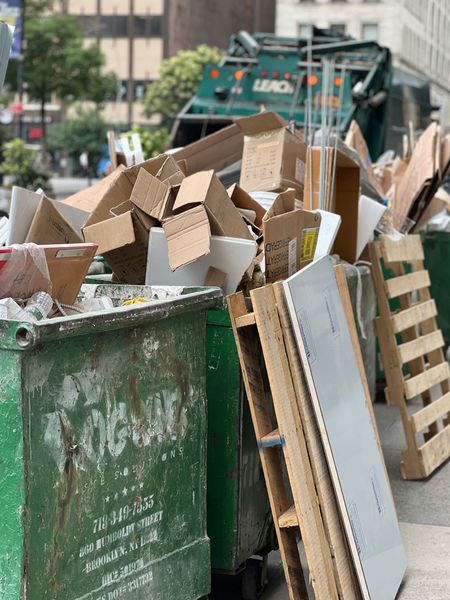 Green trash bins overflowing with cardboard boxes and wooden pallets; a garbage truck in the background.