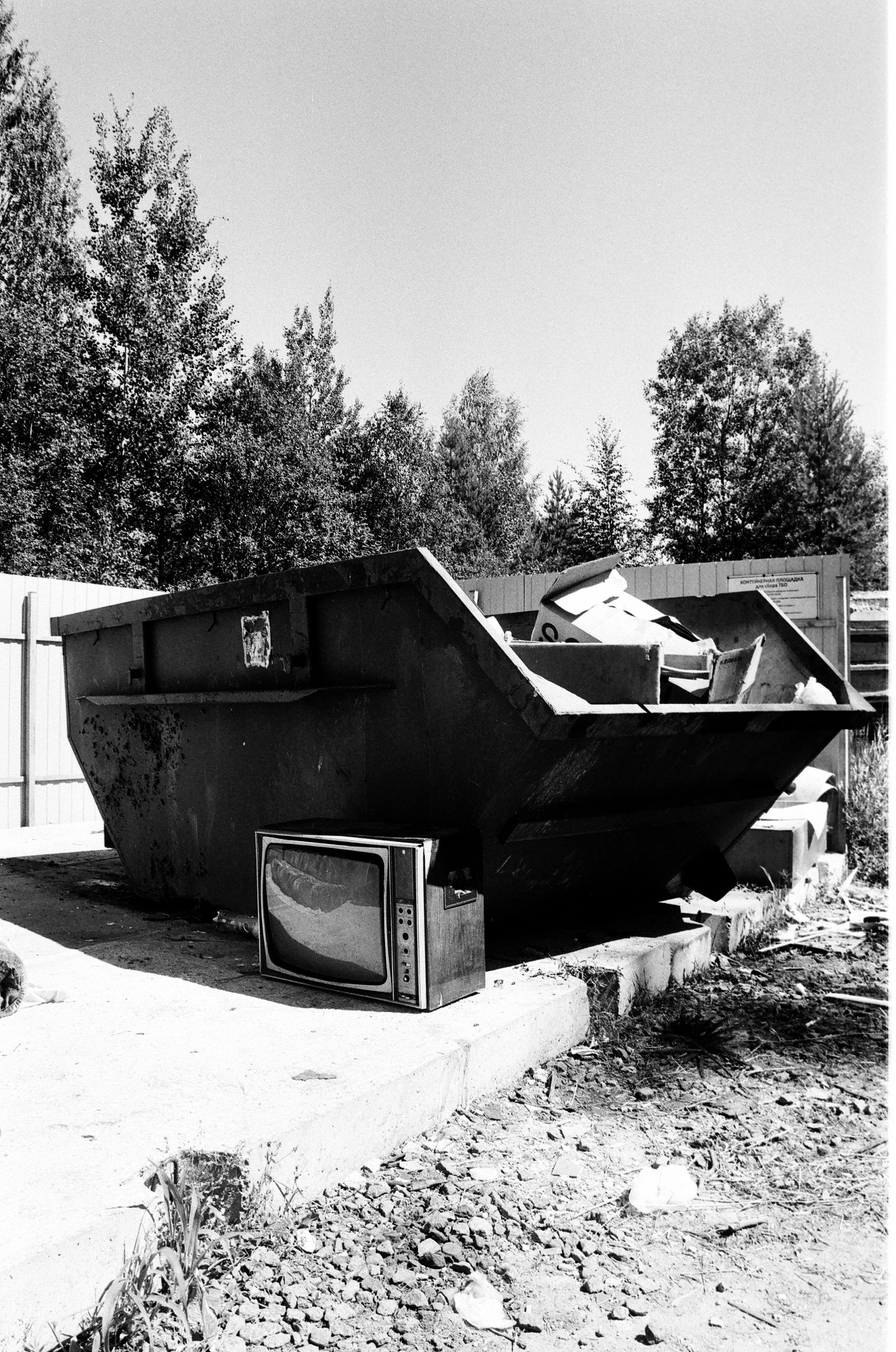 A large, open dumpster filled with trash; an old TV sits outside; trees in background.