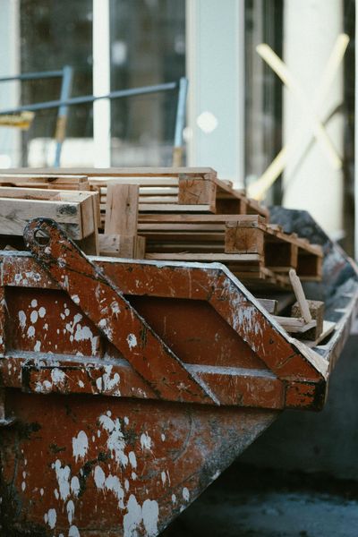 Construction dumpster filled with wooden pallets, orange with white paint splatter, building in background.
