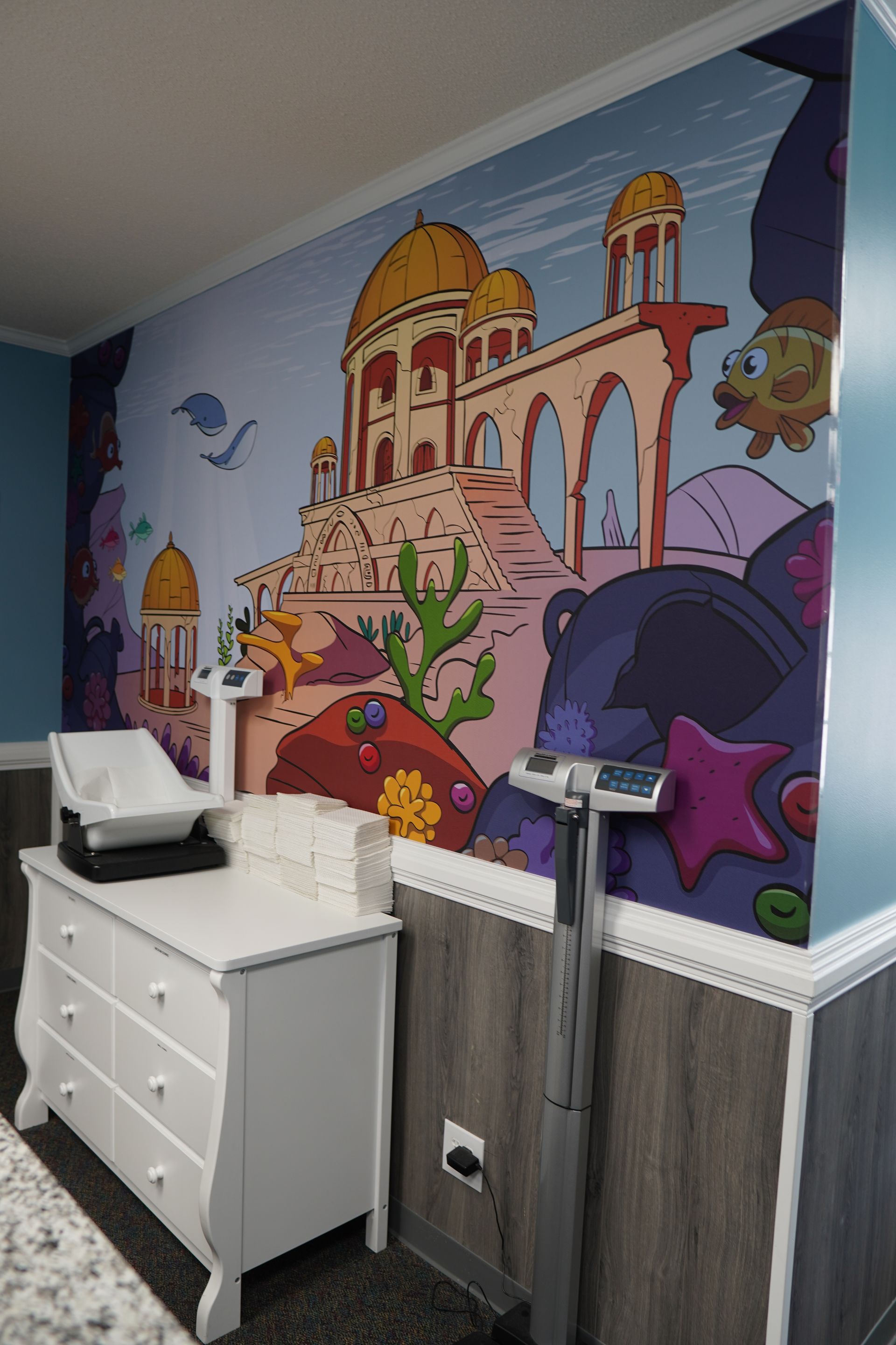 A nursery room with a colorful undersea mural, a changing table, and a weight scale.