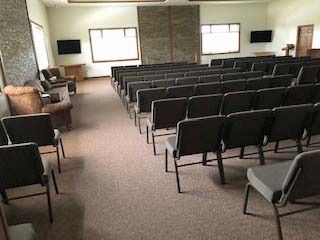 Rows of chairs in a sparsely-filled room, possibly a chapel or auditorium. Brown carpet, neutral walls, and two small TVs.