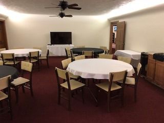 Round tables with chairs in a room, a TV on a stand, two ceiling fans.
