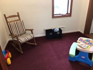 A room with a rocking chair, a toy table, a storage bin, and a window. Red carpet.