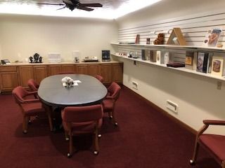 Funeral home arrangement room with oval table, chairs, display shelves, and red carpet.