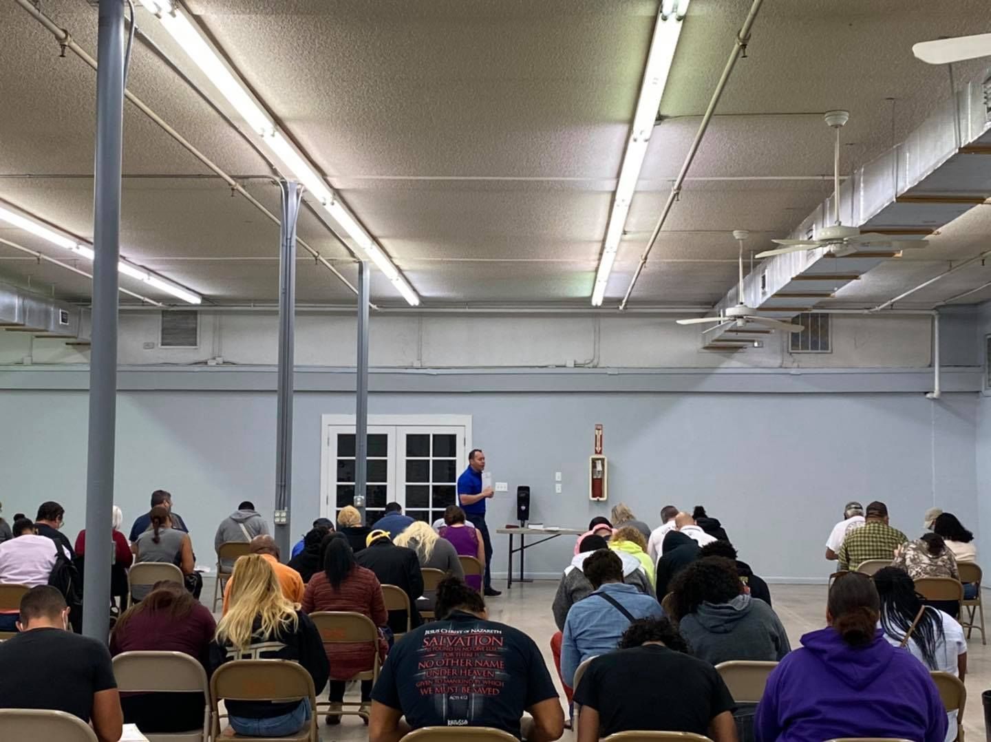 People in chairs listen to a speaker in a large room with fluorescent lights.