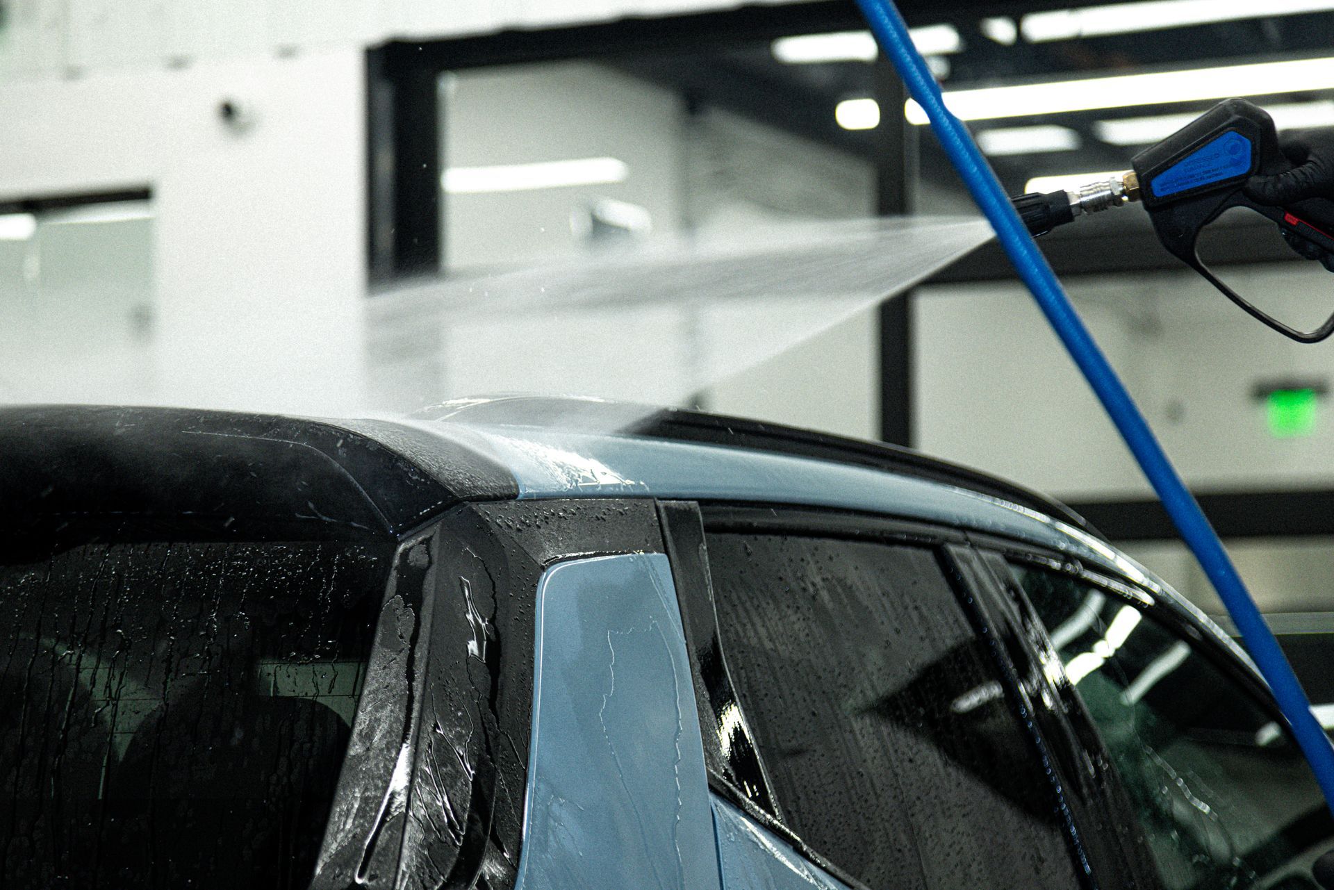 A person washing a blue car roof with a high-pressure water hose in a bright, indoor setting.