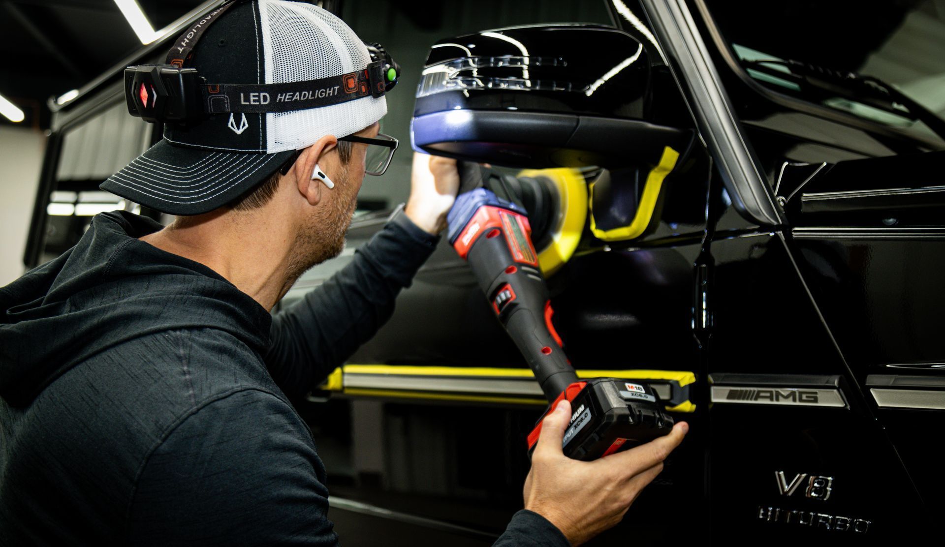 A person working on the side mirror of a black car, holding a tool with a flashlight. They wear a cap, headlamp, and earplugs.