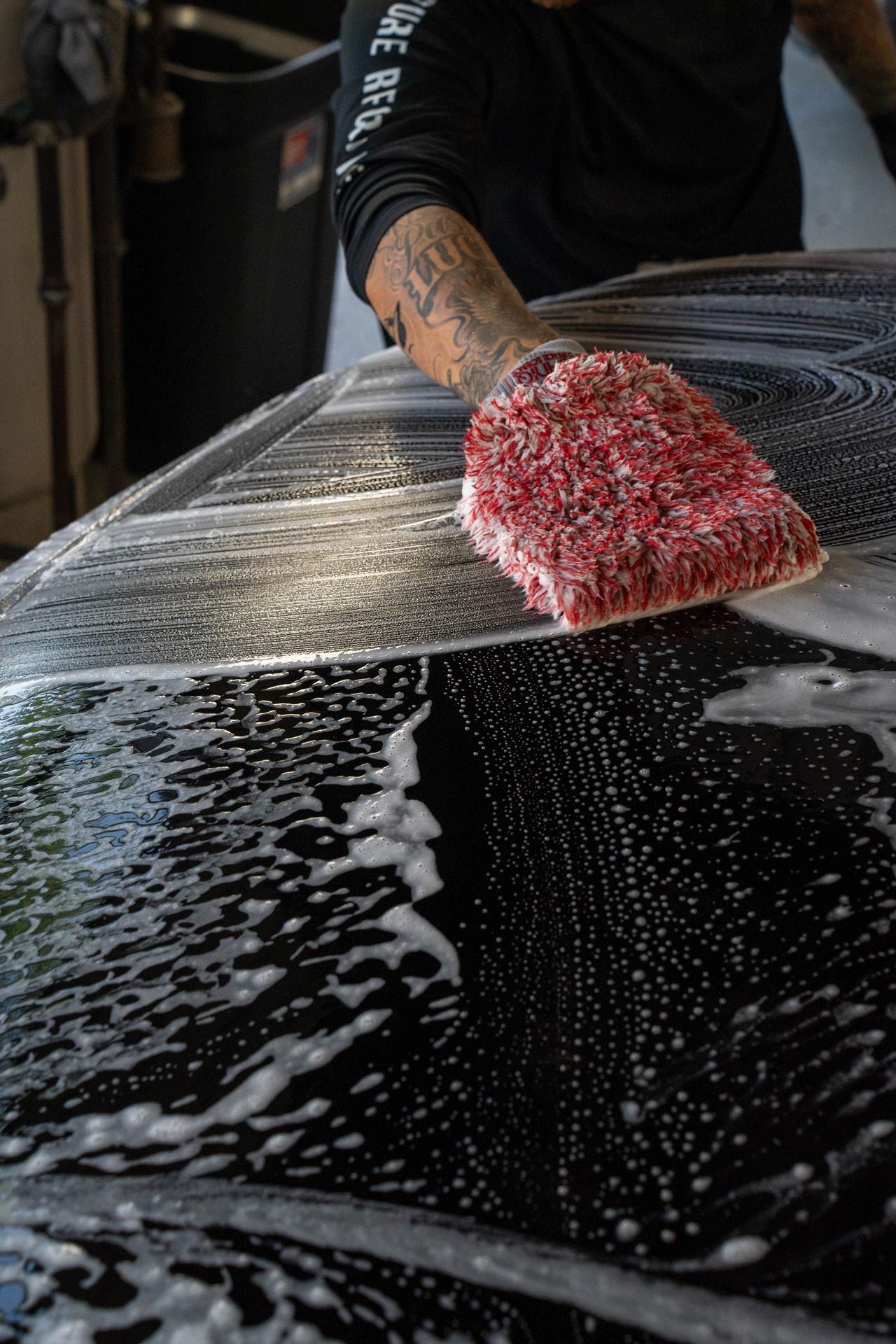 A person washing a black car with a soapy red mitt, creating a foamy surface.