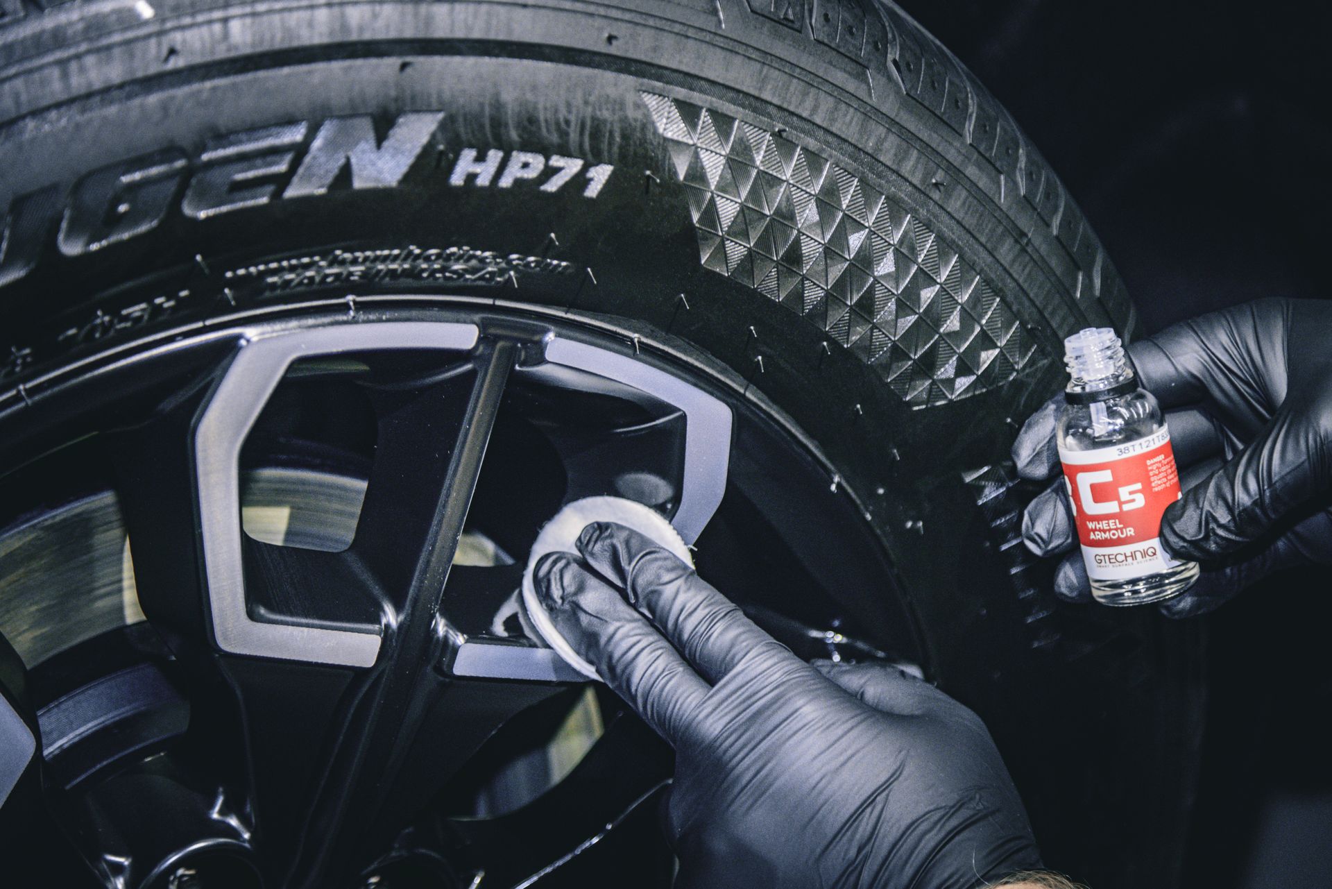 A person in black gloves applies a ceramic coating to a car rim with a white applicator, holding a bottle of C6 ceramic coating.