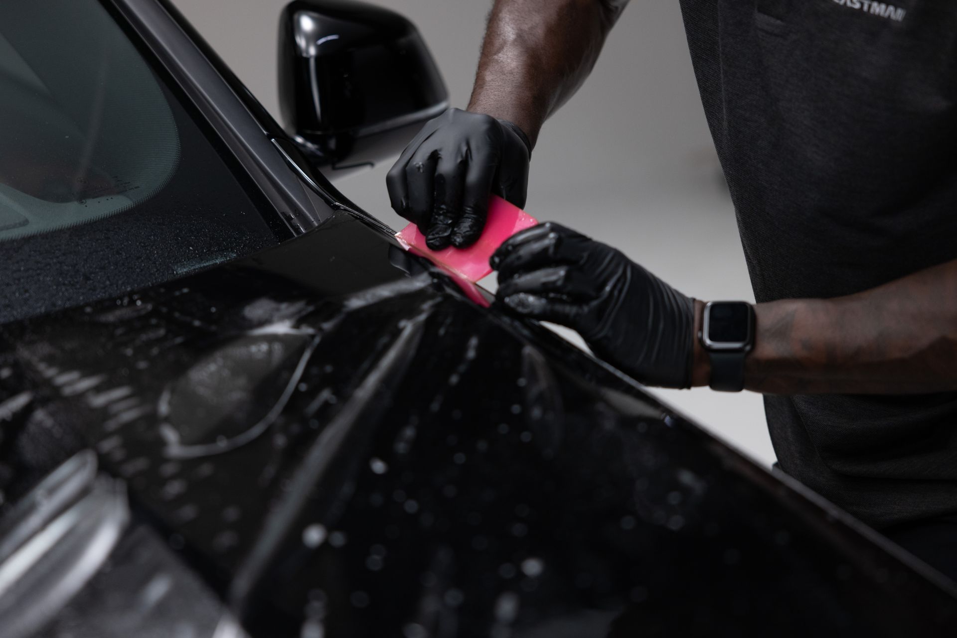Person wearing black gloves applies protective film to a black car's hood, using a pink tool.