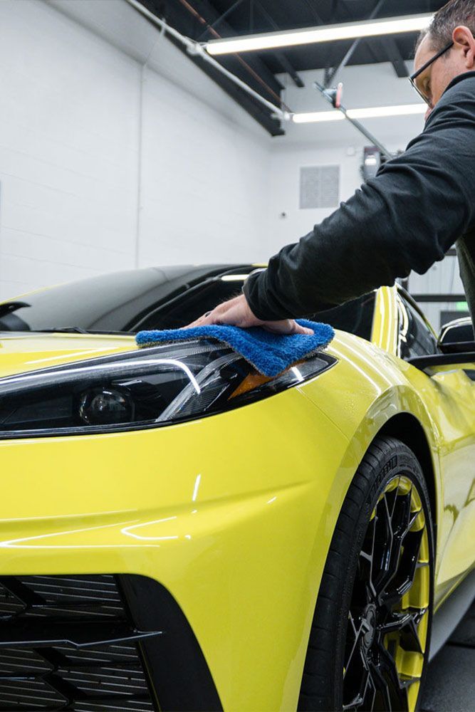Person wiping down the hood of a bright yellow sports car with a blue microfiber cloth in a well-lit garage.