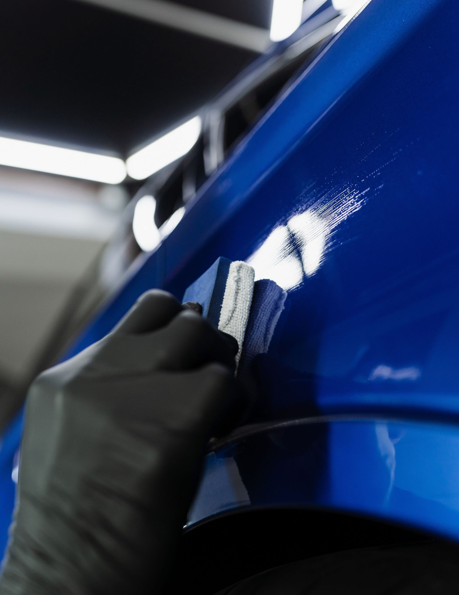 Gloved hand applying ceramic coating to a shiny blue car panel in a well-lit garage.
