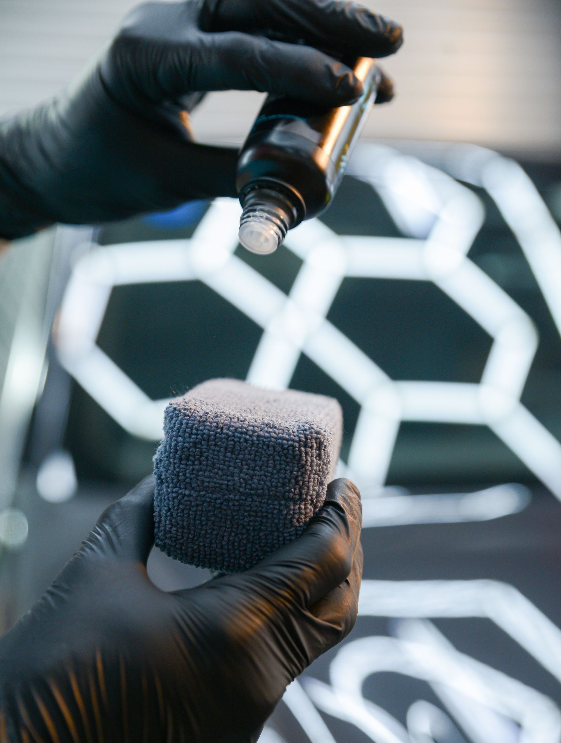 Person wearing black gloves applying car wax to a grey applicator sponge near a car.