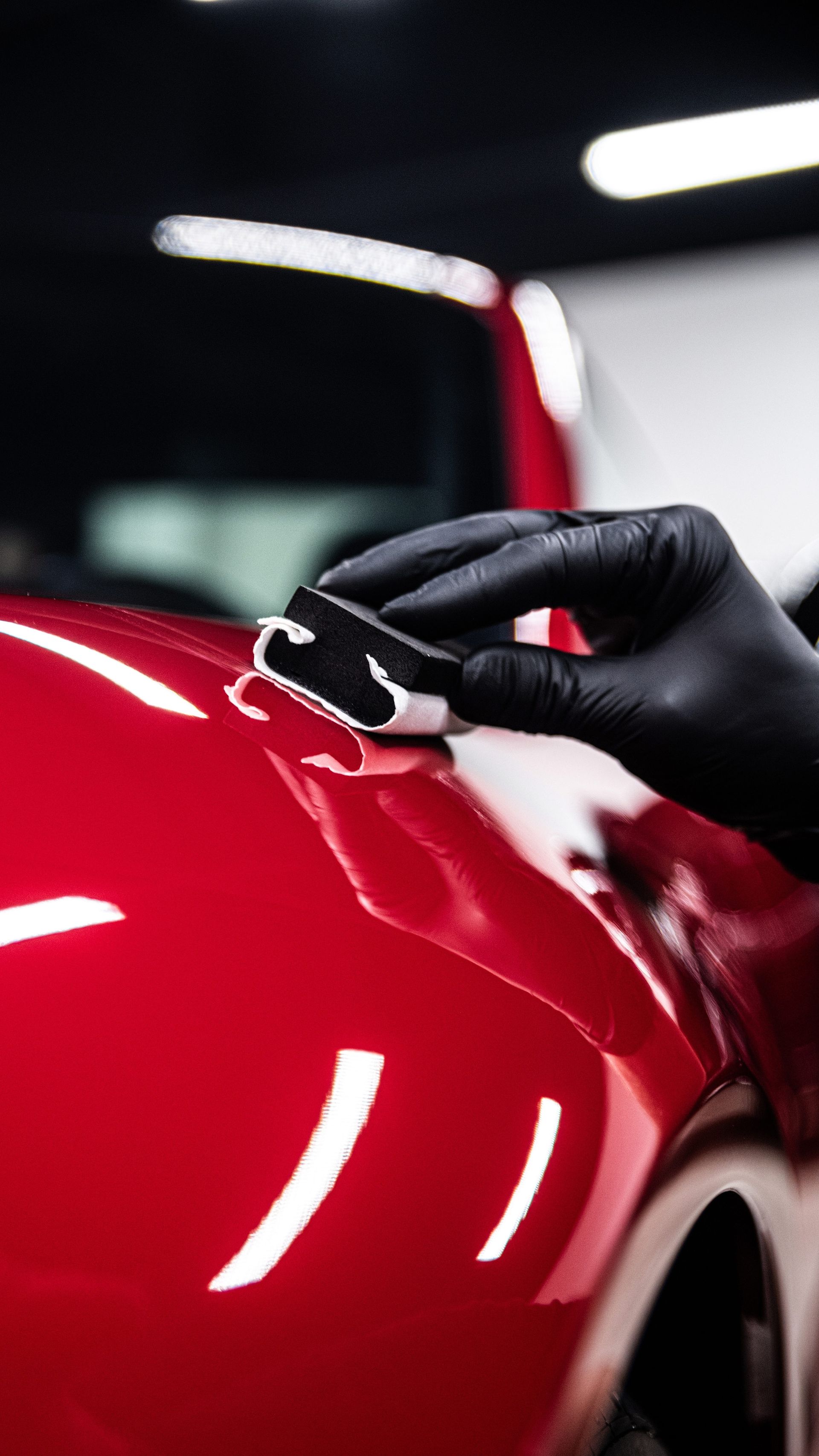 Person wearing a black glove applying a ceramic coating to a shiny red car in a garage.