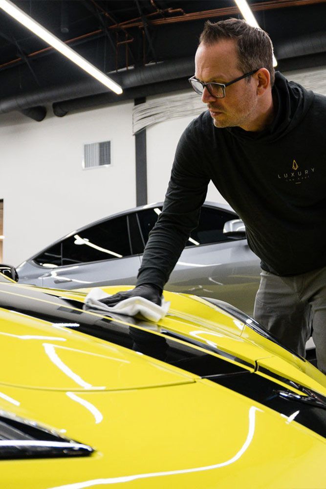 Man in glasses wipes a bright yellow car with a white cloth. He’s in a garage with another car in the background.