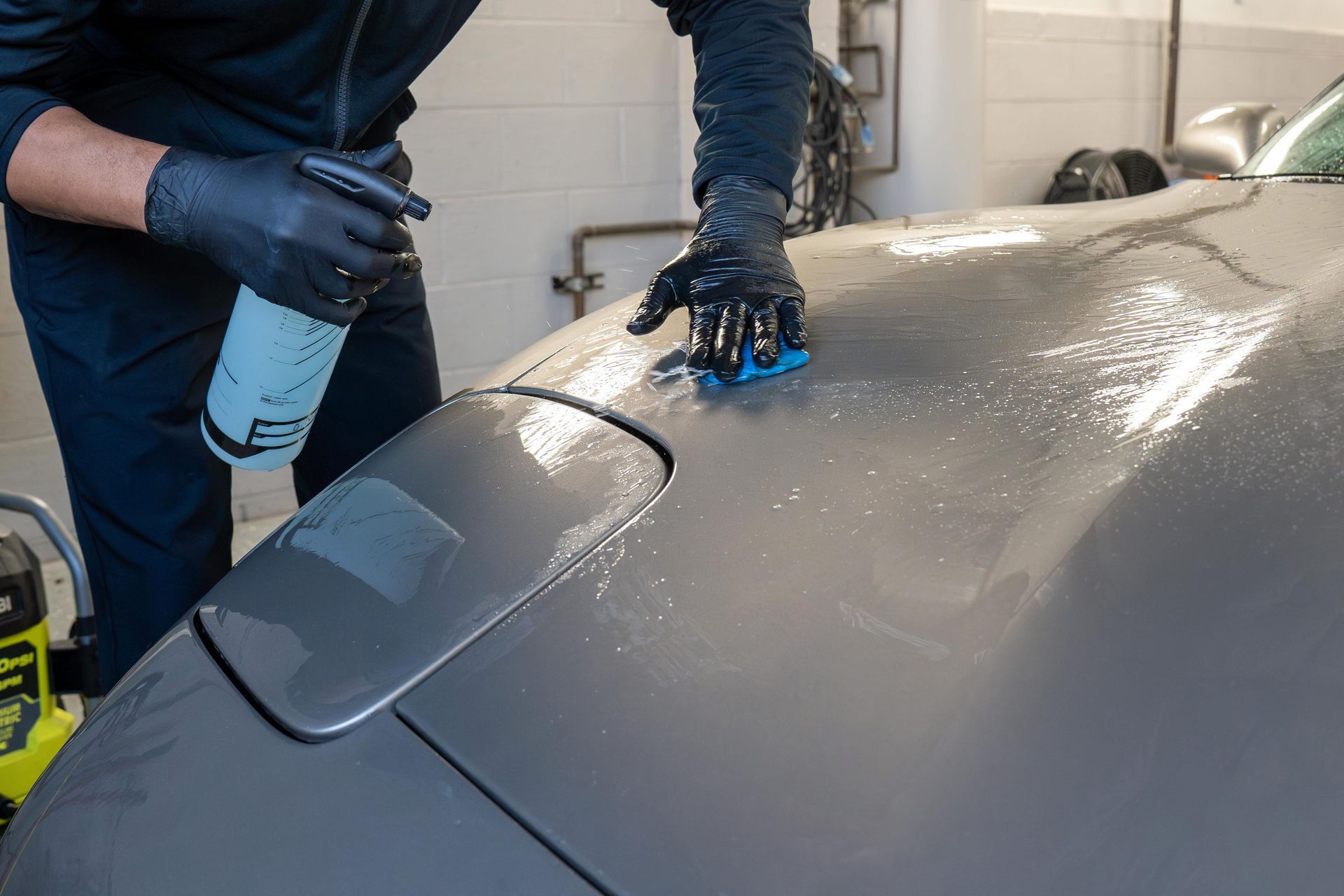 Person wearing gloves sprays and wipes a gray car hood.