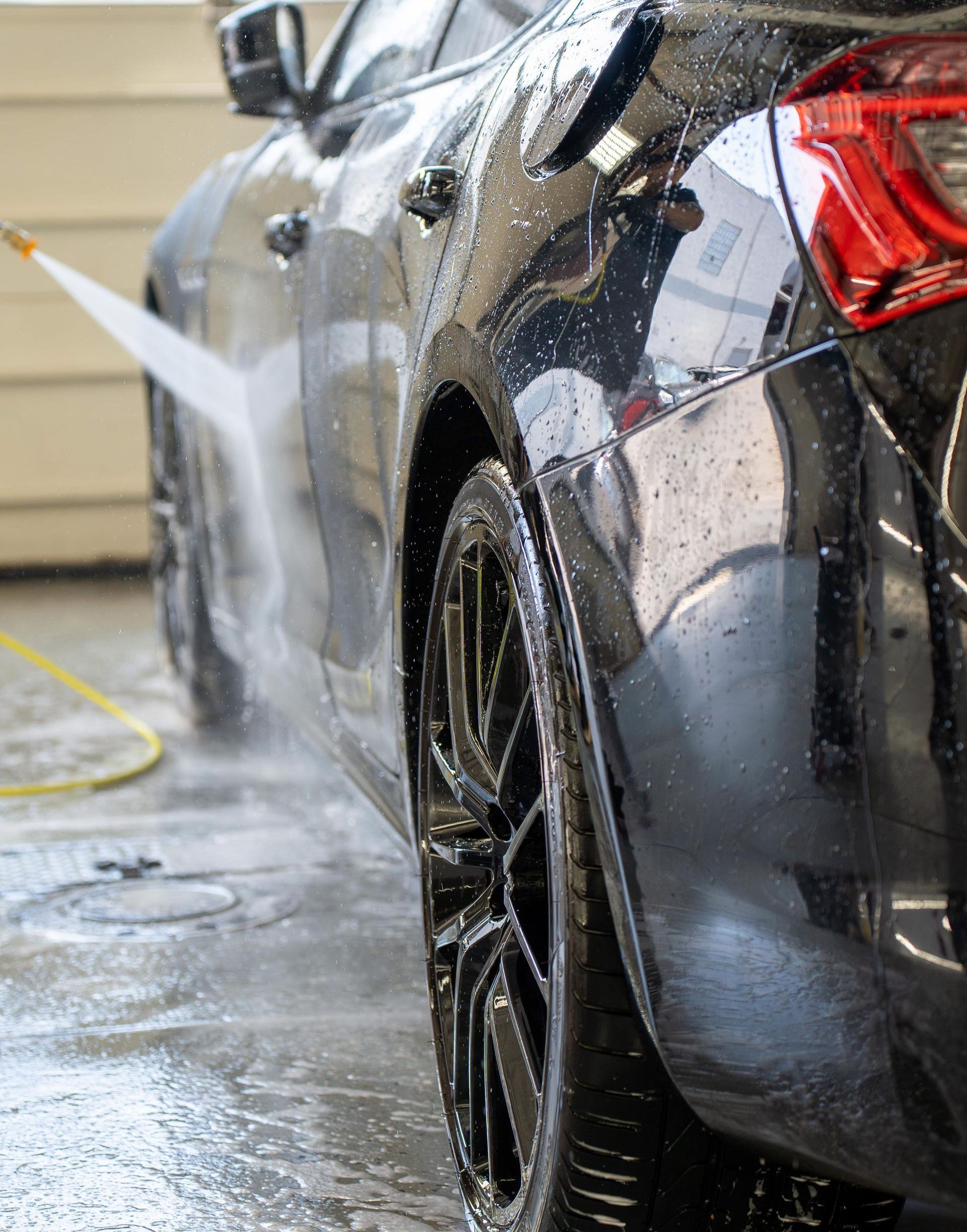 Black car being washed with a high-pressure water hose at a car wash. Close-up of the rear tire, door, and taillight.