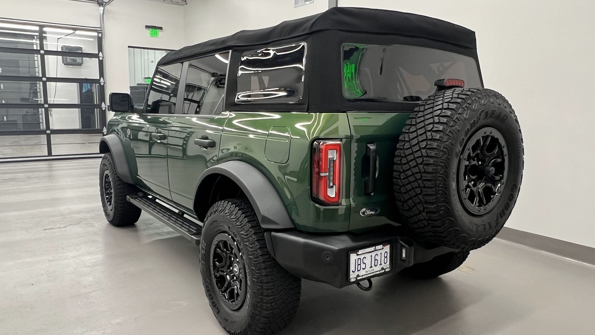Green Ford Bronco with a black soft top, black wheels, and spare tire, parked indoors.