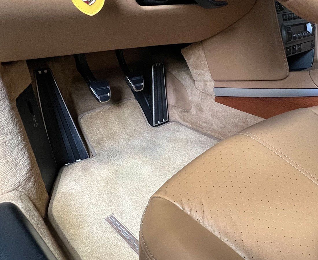 Interior car shot of a tan carpeted floor with brown leather seat, brake, accelerator, and clutch pedals visible.