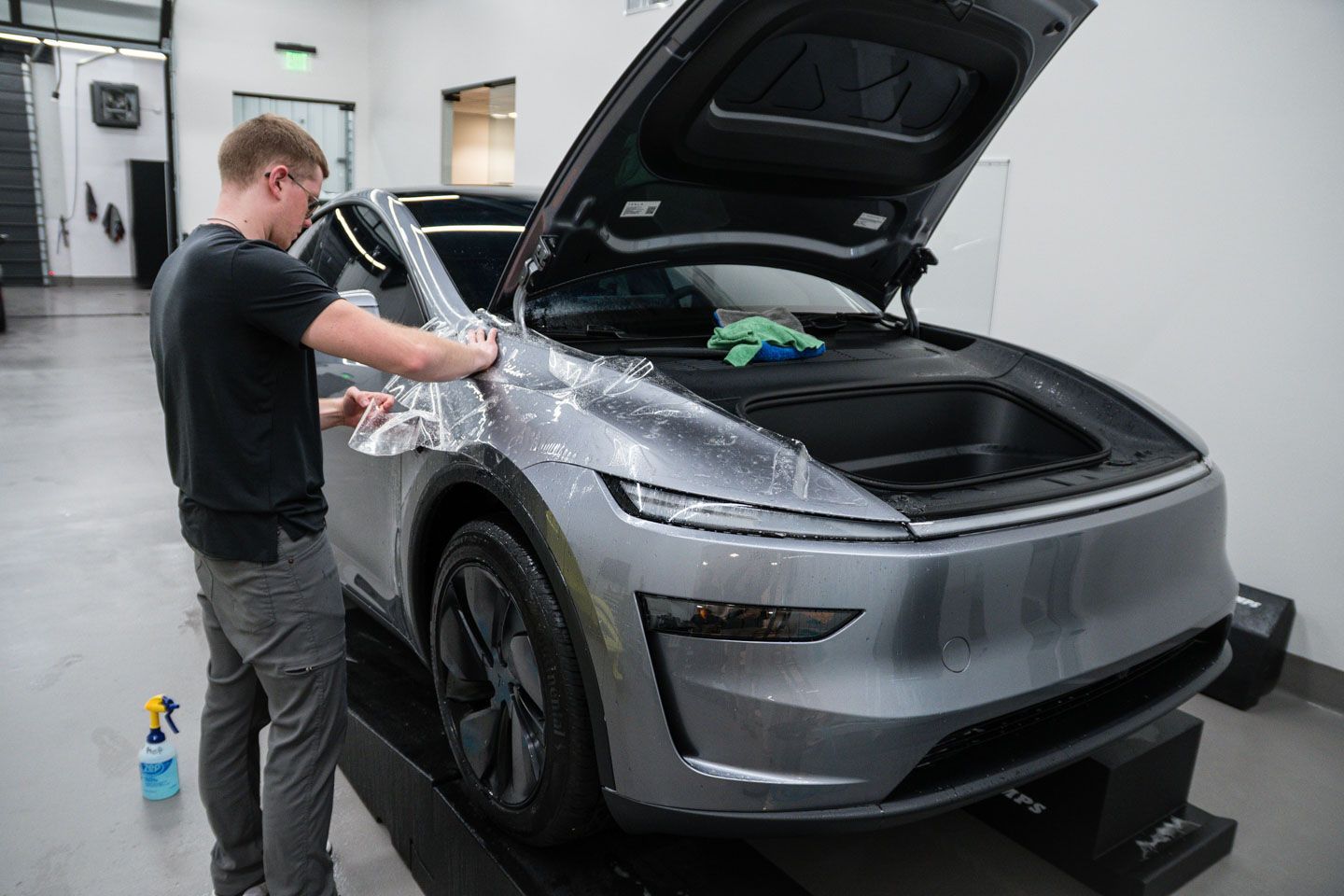 Man applying protective film to the front of a gray electric car in a bright garage. The car's hood is open.
