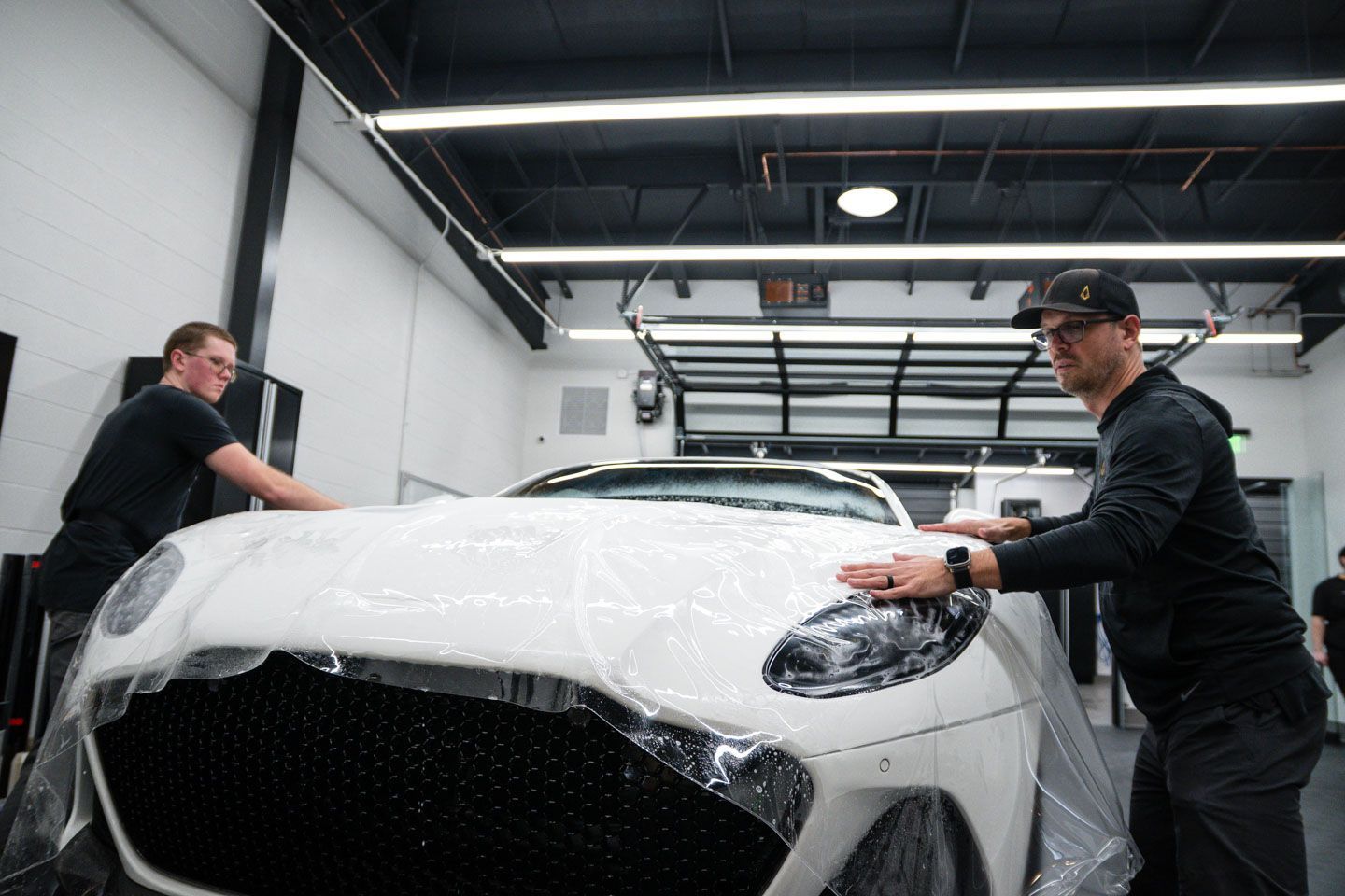 Two people wrapping a white sports car in protective plastic inside a garage.