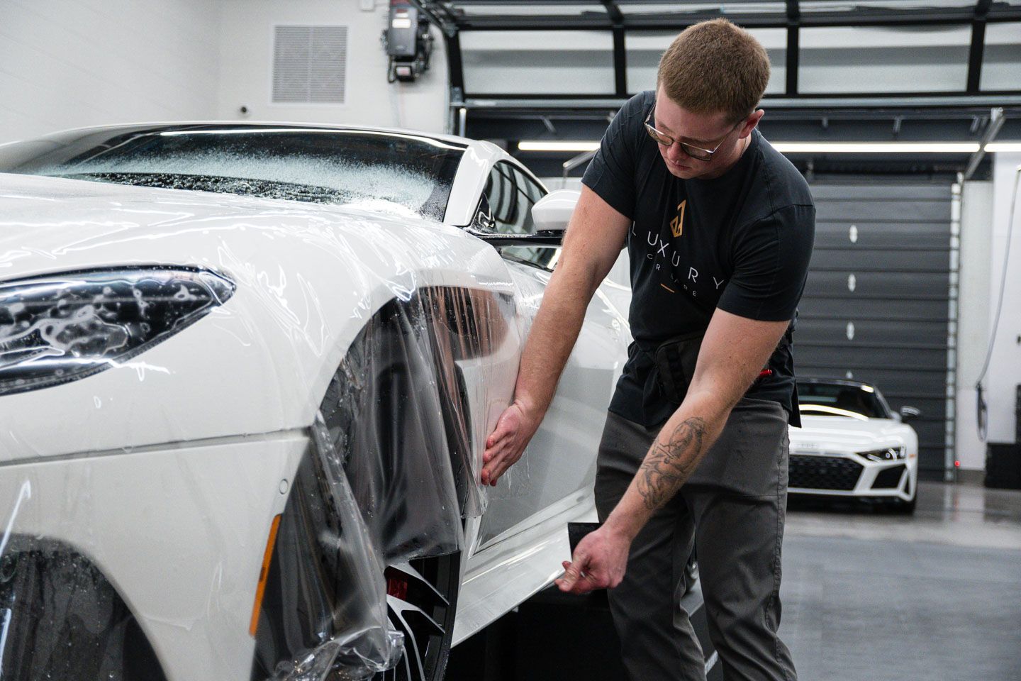 A man is applying a protective film to a white car in a garage.